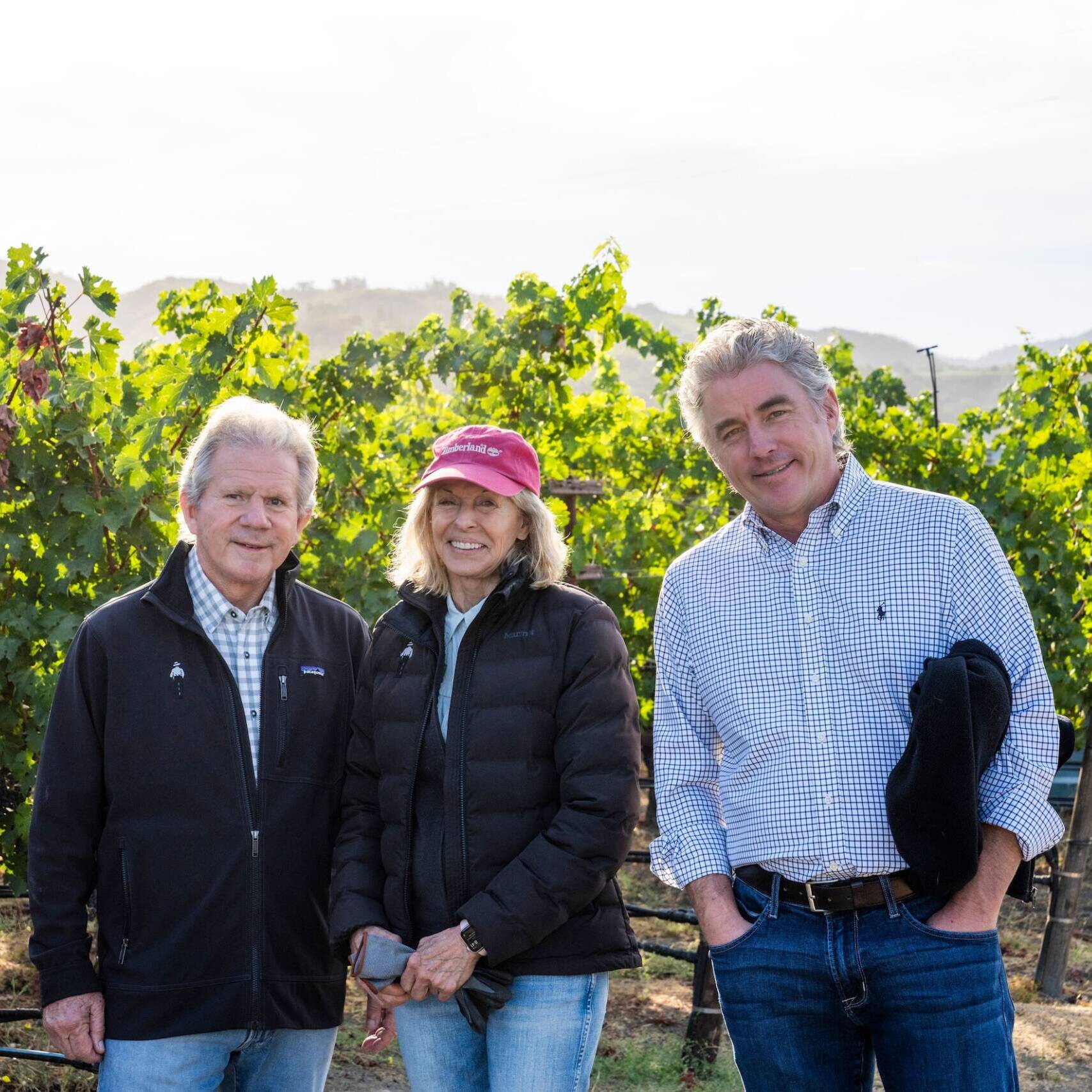 Bart, Daphne & Greg Araujo stand together outdoors in front of grapevines, all wearing jackets and smiling at the camera.